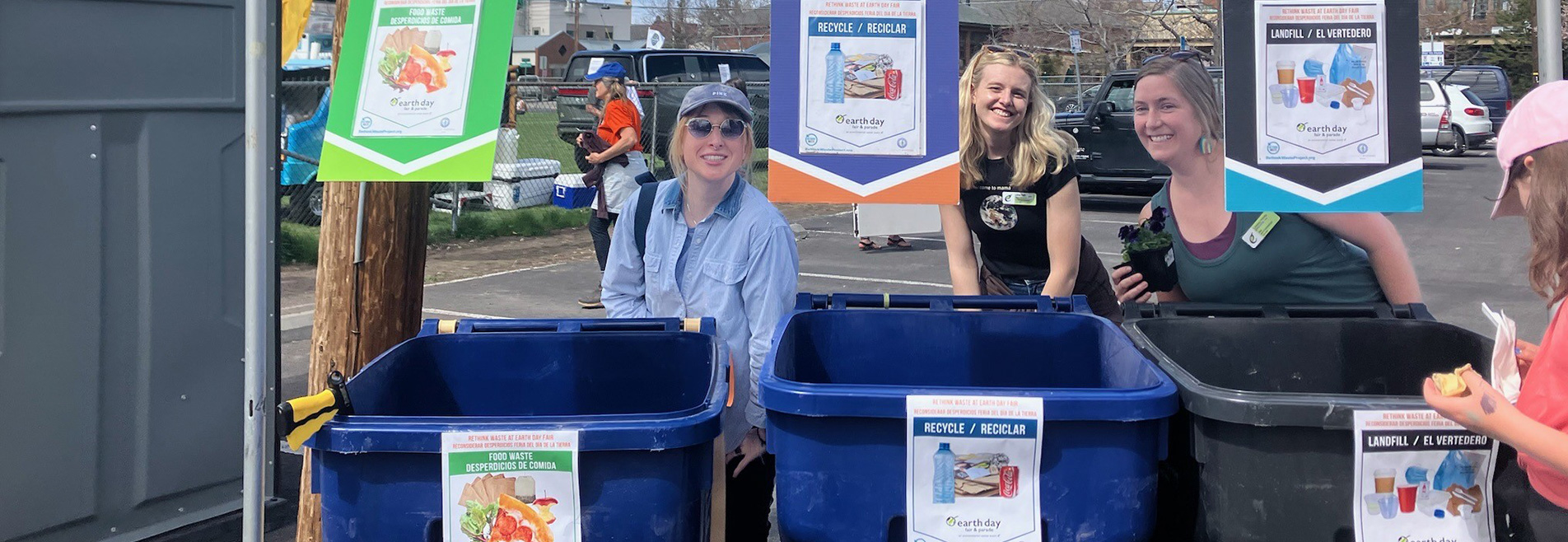 People behind recycling bins. Bins are blue and gray. Signs on each bins indicating what type of material belongs in each bin.