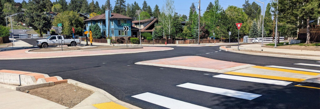 Roundabout shown from street level. Black asphalt, gray sidewalks, street signs, and houses surrounding.