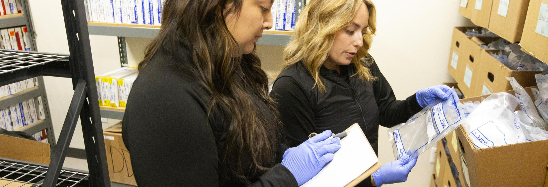 Two people examining bags of evidence from a brown box on a shelf in a room.