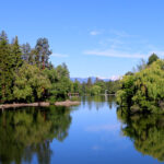 Mirror Pond with Drake Park to the left. Water is like glass reflecting the landscape and mountains on a blue sky day.