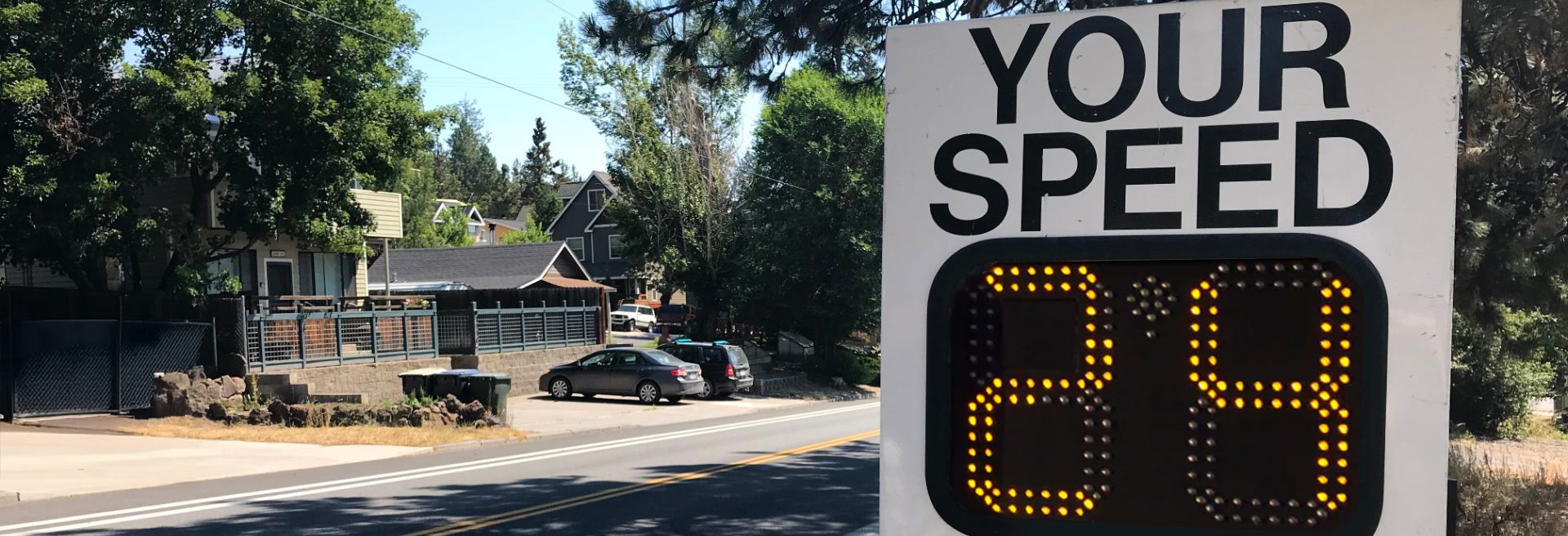 Speed radar in a neighborhood with houses and trees. Radar is a white sign with digital numbers indicating driver's speed.