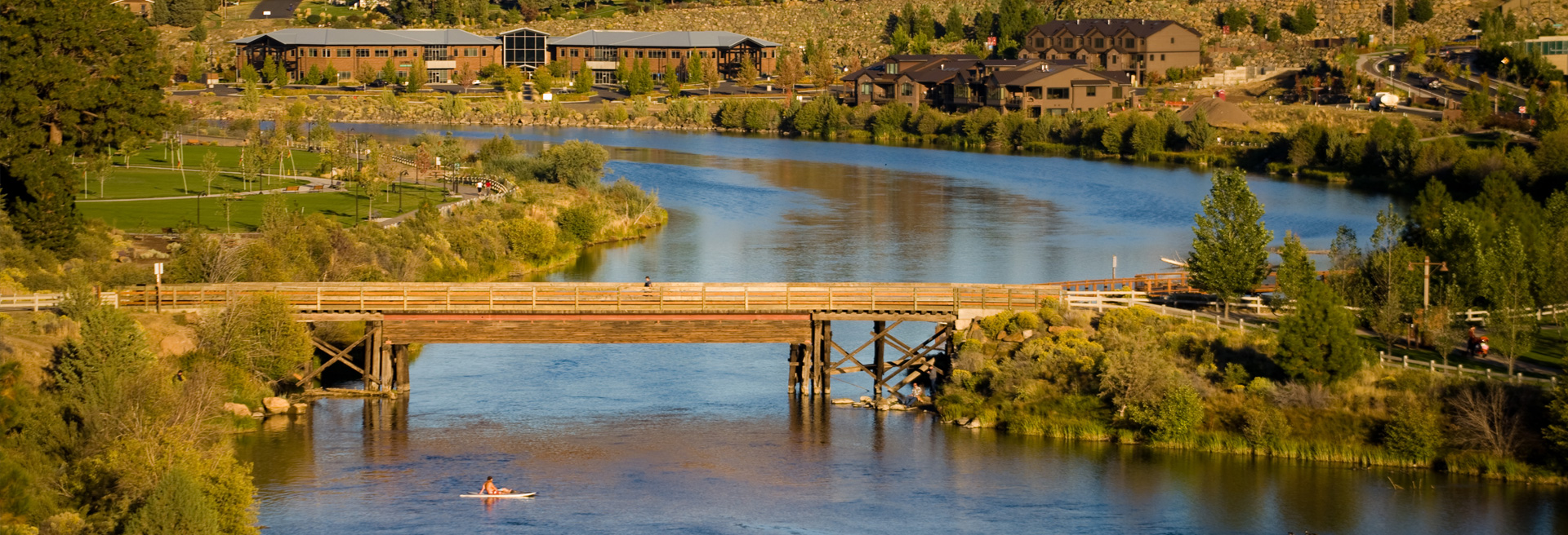 Image of footbridge over the Deschutes River. Green grass and trees and Farewell Bend park border the river.