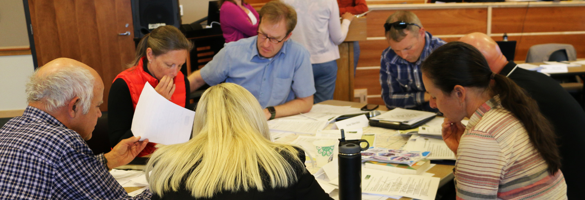 Image of people working around a table. Men and women, torsos up, papers on table.