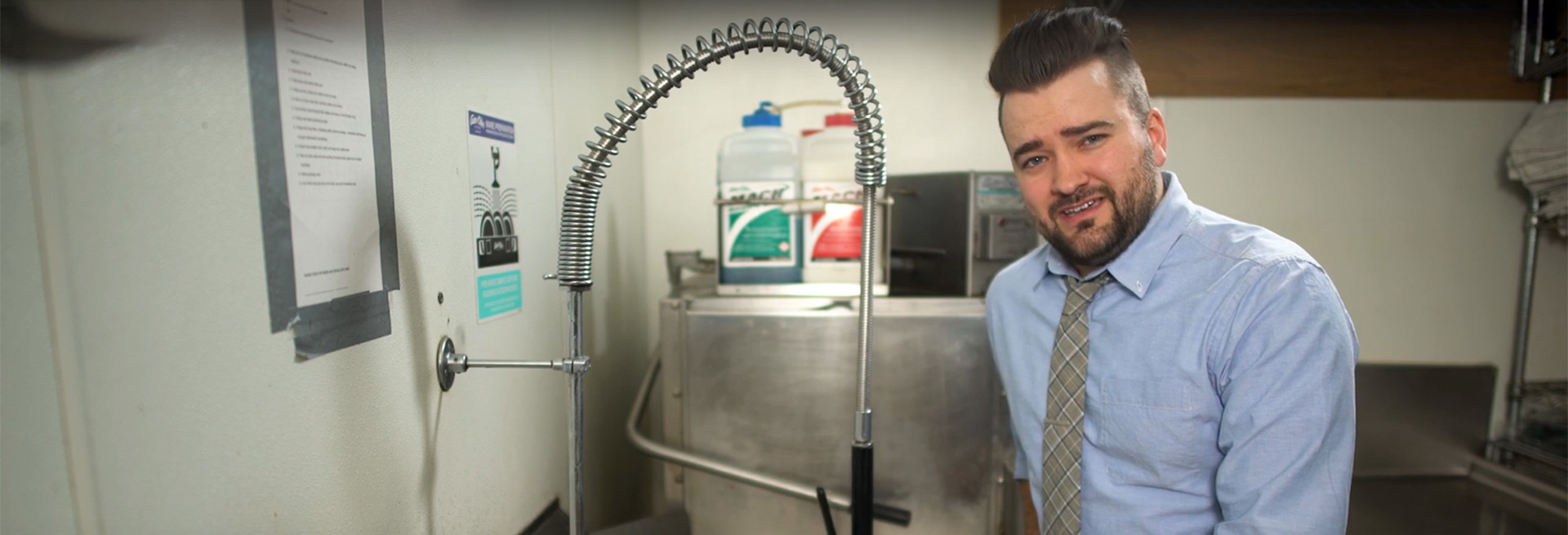 Man with brown hair and cropped beard standing at a sink in a commercial kitchen. He is wearing a gray tie and light-blue shirt.