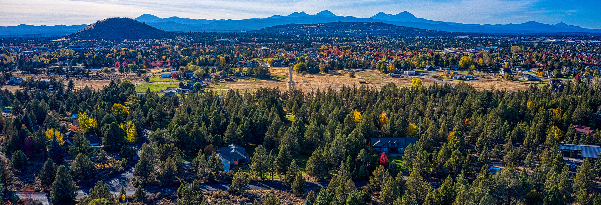 Drone shot of Bend looking northeast. Pilot Butte in the mid-ground. Party cloudy skies. Trees, roads, and houses.