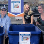 People behind recycling bins. Bins are blue and gray. Signs on each bins indicating what type of material belongs in each bin.