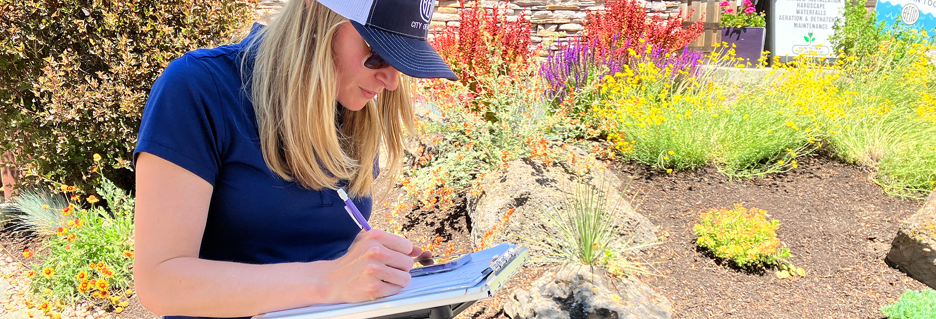 Female wearing a blue shirt and blue baseball cap. She is wearning sunglasses and writing on a clipboard.