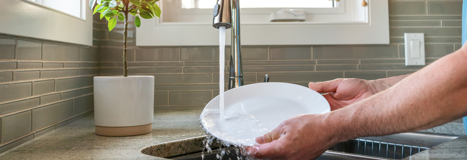Hands holding a white plate being rinsed by water from a kitchen faucet.