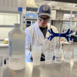 Man in a lab coat wearing a hat uses lab equipment in a scientific laboratory.