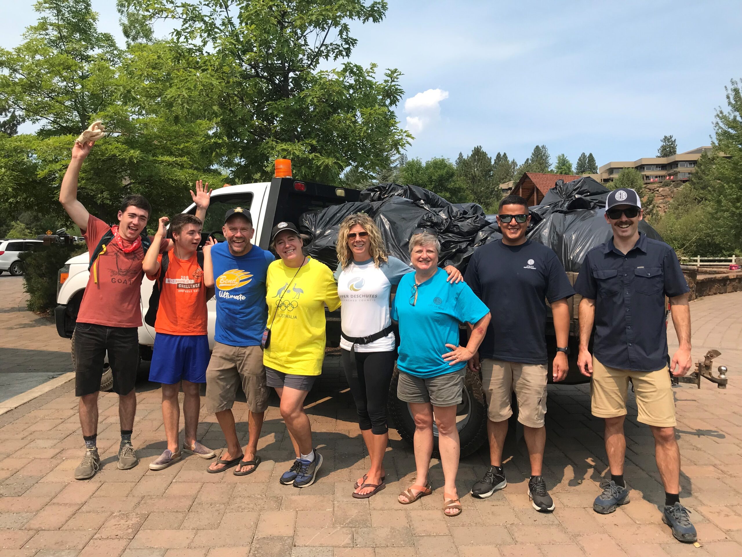 A group of smiling people pose in front of a truck full of garbage bags