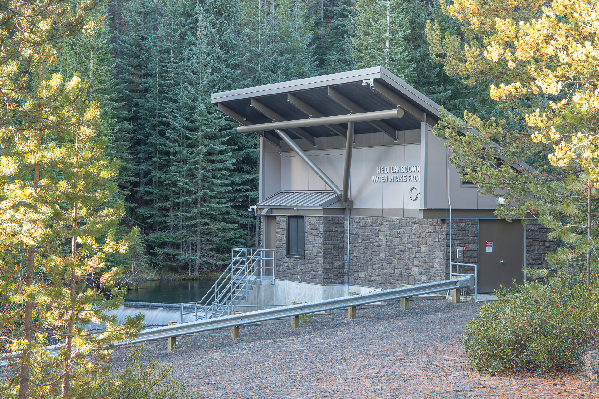 A small building with a slated roof site in the forest next to a stream and small waterworks.