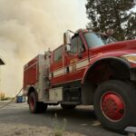 Camión de bomberos frente a una casa, con humo al fondo.