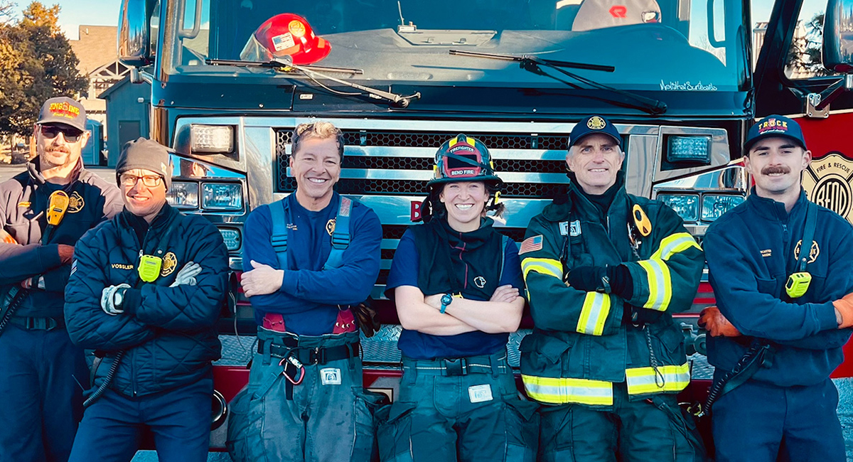 Group of people wearing fire fighting gear, standing in front of a fire engine.