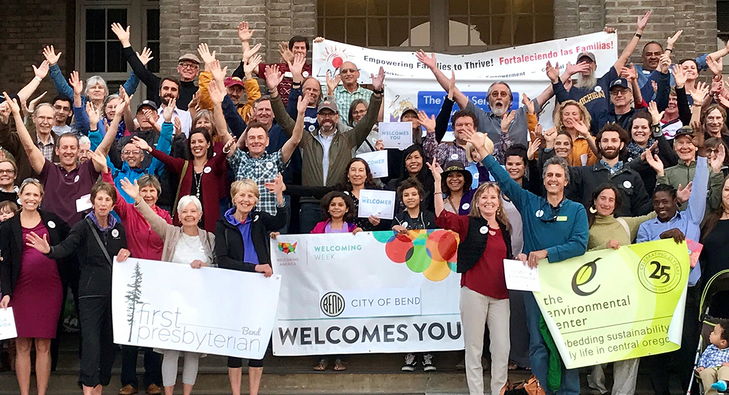 Diverse group of people standing on steps with their arms open and upraised as a sign of welcoming.