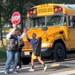 School bus with as crossing guard and a child walking in front of the stopped bus.